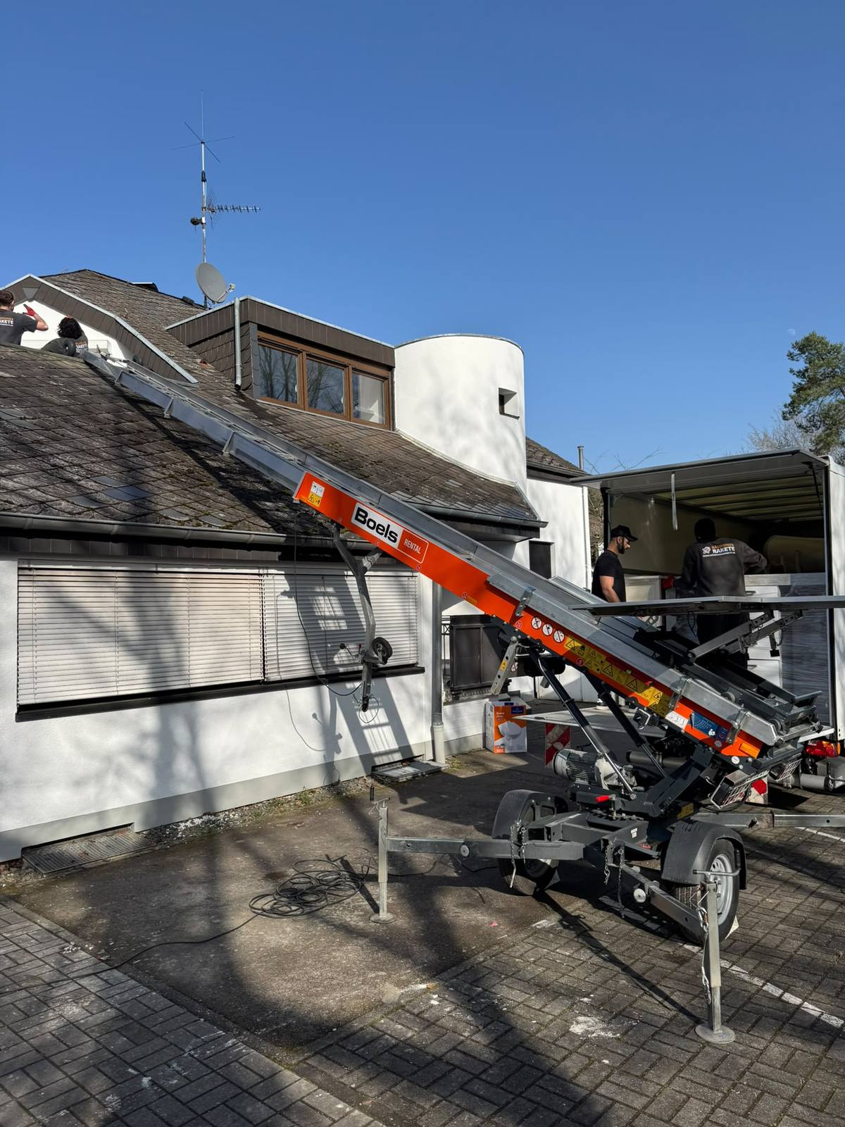 Möbellift im Einsatz an einem Dachgeschoss mit zwei Mitarbeitern von RäumungsRakete auf dem Dach, im Hintergrund ein geöffneter Umzugswagen bei strahlendem Sonnenschein