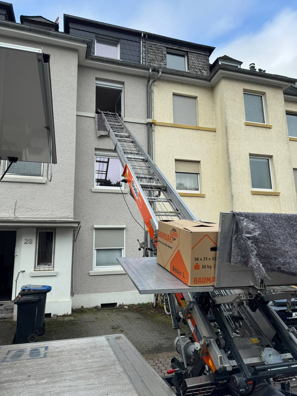 Nahaufnahme der Lift-Plattform mit einem Umzugskarton und einem eingerollten Teppich, im Hintergrund das geöffnete Fenster im Obergeschoss eines Wohnhauses