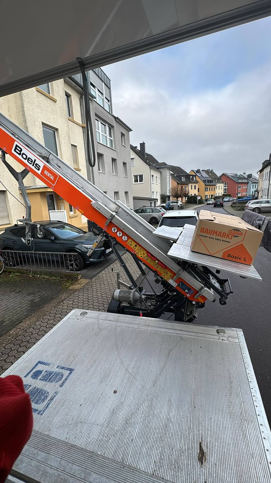 Blick aus dem geöffneten Umzugswagen auf den orangefarbenen Hubarm des Möbellifts mit einem Umzugskarton der Marke Globus Baumarkt auf der Transportplattform, im Hintergrund eine Trierer Wohnstraße